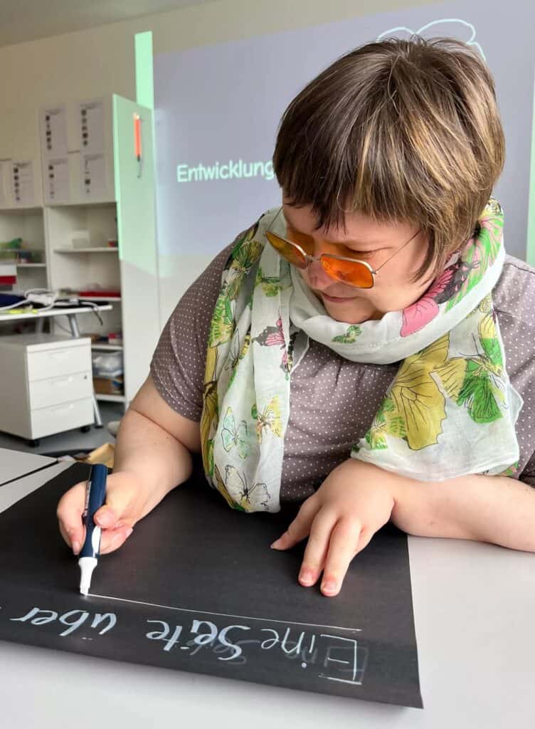 Ellen Keune, die mit einem weißen Stift auf ein schwarzes Papier an einem Tisch schreibt. Sie schreibt: eine Seite über