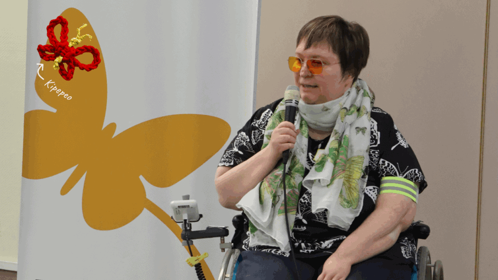 Ellen Keune spricht in ein Mikrofon. Sie trägt ein schwarzes T-shirt mit weißen Schmetterlingen und einen bunten Schal mit Schmetterlingen. Hinter ist ein Banner mit einem Schmetterlings-Logo